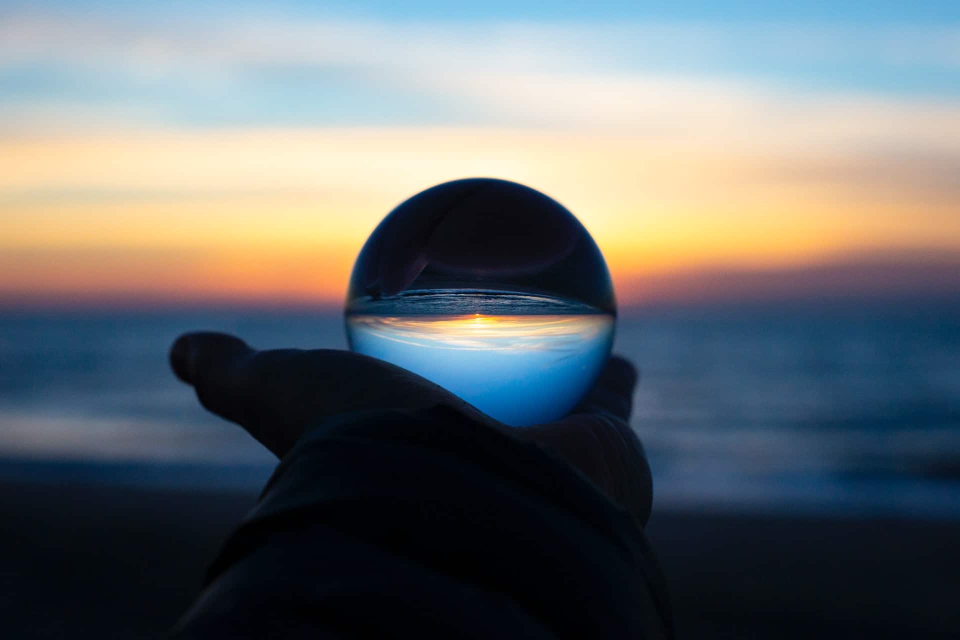 A person holding a glass ball in front of the ocean at sunset.